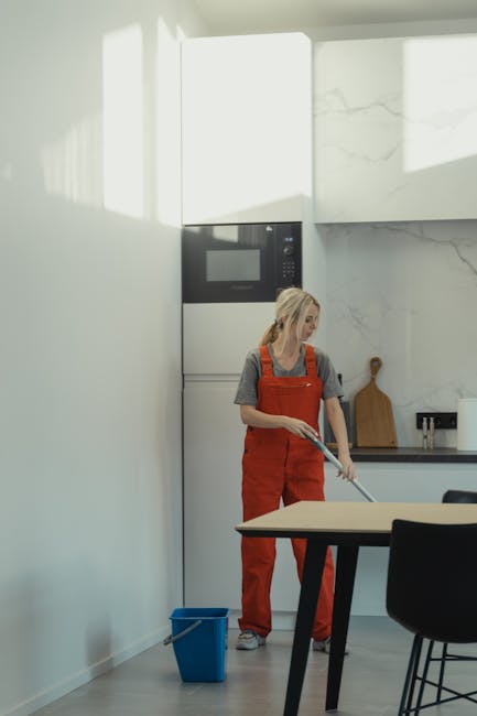 A young woman with dark hair, wearing a grey t-shirt and bright orange overalls, is standing inside a modern kitchen. She is holding a vacuum cleaner hose, which is connected to a yellow floor cleanin